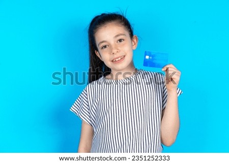 Close up photo of optimistic Beautiful caucasian kid girl standing over blue studio background hold card