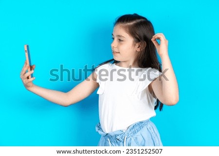 Beautiful caucasian kid girl standing over blue studio background smiling and taking a selfie ready to post it on her social media.