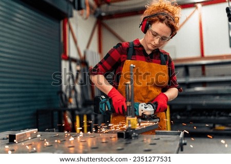 A young girl who is an apprentice in a metal workshop uses a grinder and works on a metal bar, she is wearing protective equipment