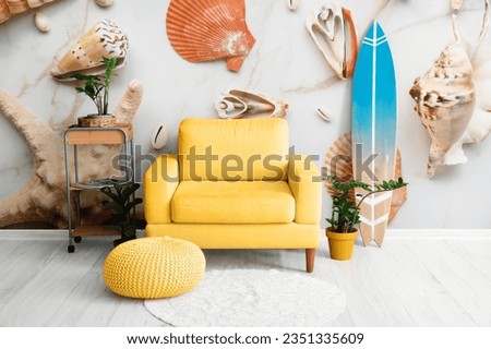 Wooden surfboard with yellow armchair and houseplants near wall with printed sea shells and starfishes