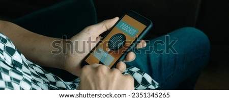 man, in casual wear, sitting in a chair, listening a mock podcast in his smartphone, that reads the text podcast in its screen, in a panoramic format to use as web banner Royalty-Free Stock Photo #2351345265