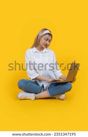 Working on laptop, full length portrait of attractive young blonde woman sitting on floor working on laptop. Holding pc notebook computer. Isolated yellow background studio. Cute blonde girl in 20s. 
