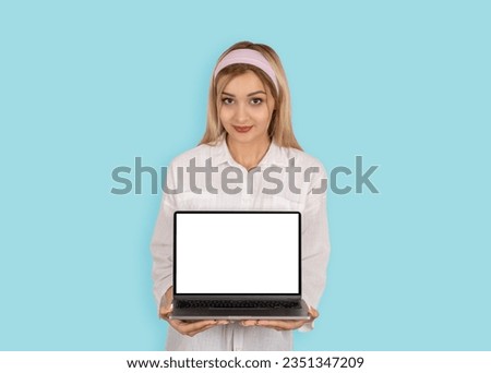 Laptop mock up, pretty cute blonde girl in 20s wearing white shirt holding laptop mock up. Standing over light blue pastel studio background. Empty blank screen pc computer notebook. Lifestyle concept