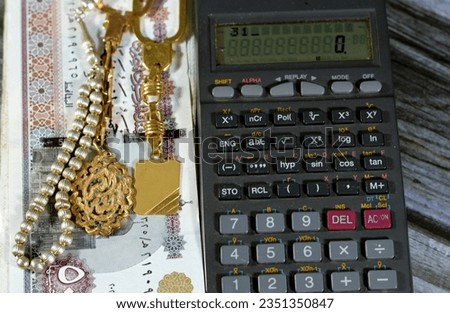 A calculator, silver rosary and gold keyring medals with a text Allahu Akbar, translation (God is the greatest), on pile of Egyptian 50 LE EGP pounds, Egypt money exchange rate with gold and silver