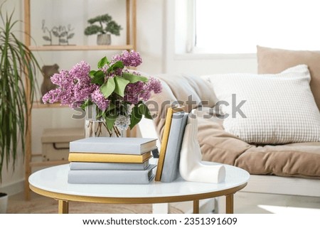 Stylish holder for books with vase of beautiful lilac flowers on coffee table in living room