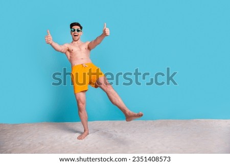 Full size photo of attractive young man showing thumbs up dressed stylish yellow shorts isolated on blue color background summer vacation