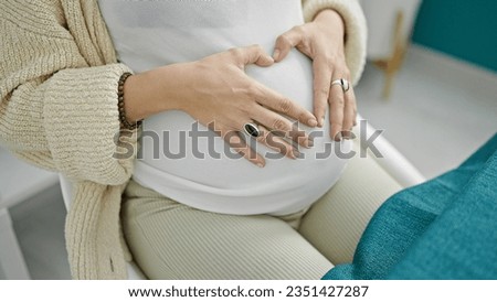 Young pregnant woman sitting on table doing heart symbol with hands on belly at dinning room