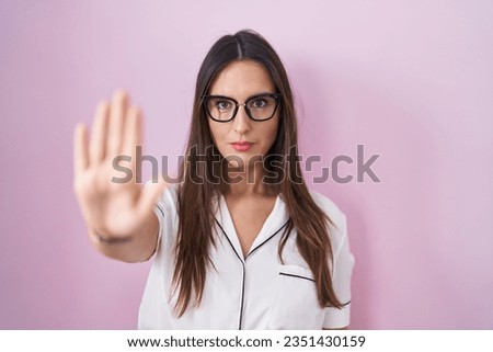 Young brunette woman wearing glasses standing over pink background doing stop sing with palm of the hand. warning expression with negative and serious gesture on the face. 