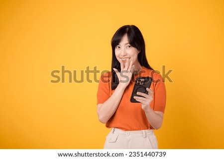 Curious Asian woman 30s, wearing orange shirt, using smartphone with fist up hand sign on vibrant yellow background. Unveiling new app wonder. Royalty-Free Stock Photo #2351440739