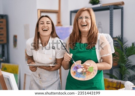 Hispanic mother and daughter painting at paint studio smiling and laughing hard out loud because funny crazy joke. 