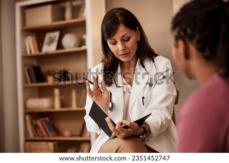 A female healthcare worker in a white uniform reads from notes while talking with an unrecognizable patient. Royalty-Free Stock Photo #2351452747