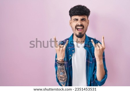 Young hispanic man with beard standing over pink background shouting with crazy expression doing rock symbol with hands up. music star. heavy concept. 