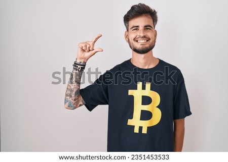 Young hispanic man with tattoos wearing bitcoin t shirt smiling and confident gesturing with hand doing small size sign with fingers looking and the camera. measure concept. 