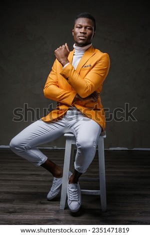 Fashionably dressed dark-skinned man posing seated on a chair against a dark background wearing a yellow jacket Royalty-Free Stock Photo #2351471819