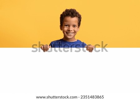 Portrait of smiling preteen black boy hiding behind blank white placard, happy kid demonstrating copy space for your text or design, showing horizontal empty board, yellow background, mockup