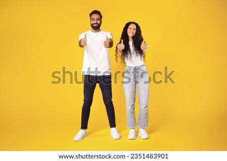 Happy arab millennial man and woman in casual showing thumb ups and smiling at camera, posing full length, showing like gestures with both hands standing over yellow background, studio shot Royalty-Free Stock Photo #2351483901