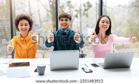 Cheerful three multiracial students, sitting at table with laptops in modern university audience, looking at camera and showing the ok sign thumbs up