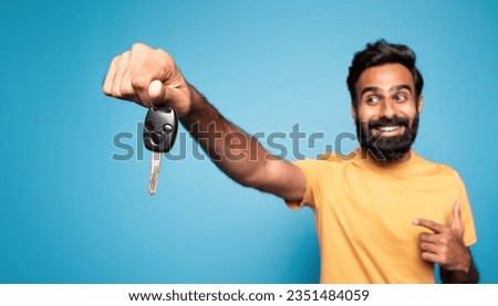 New car. Happy indian man showing auto key and pointing at it in excitement, posing on blue background, selective focus, panorama. Dreams come true, own automobile concept Royalty-Free Stock Photo #2351484059