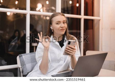 Smiling young woman hold mobile phone and show ok sign, making all right hand gesture sitting in cafe outdoors, working on laptop, wireless headphones in ears. Agreement, approval, confirm. 