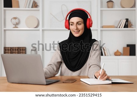 Muslim woman in headphones writing notes near laptop at wooden table in room