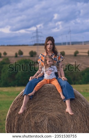 Young Caucasian Girl in jeans and colorful blouse sits with her daughter on a round bale of straw on the field.Yong mom and daughter posing on a fieldagainst cloudy sky and windmills