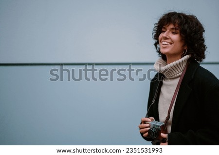 Cropped shot of a beautiful dark haired curly joyful girl standing against the gray background and looking into the distance with a big smile on her face. Holding an old retro camera. Copy space.