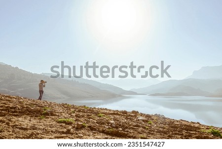 The lake under mountain landscape photographer. Unrecognizable far man taking photograph in Bouzegza Keddara,  Boudouaou, Algeria. Brown soil, bush and shiny sunlight in a blue sky reflected in water.