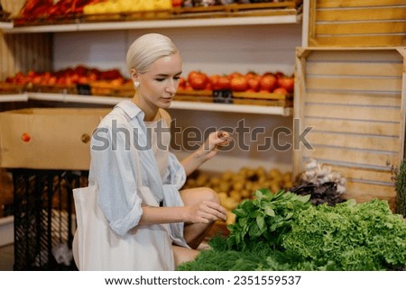 Woman is browsing and selecting various greens at a grocery store. She is wearing a light blue shirt and holding a white tote bag. Variety of healthy food options available at the store