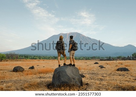 Two men, of European appearance, travelers, with backpacks on their backs, stand on a huge stone and admire the mountain at sunset, view from the back. Royalty-Free Stock Photo #2351577303