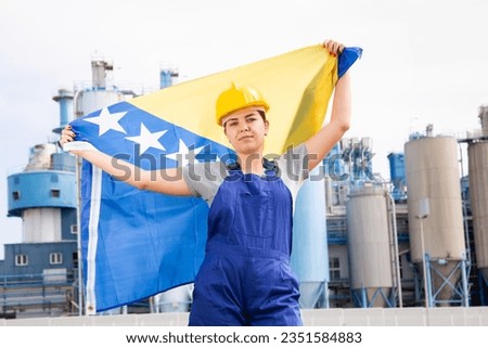 Sad female worker in hardhat with bosnian flag standing in front of factory Royalty-Free Stock Photo #2351584883