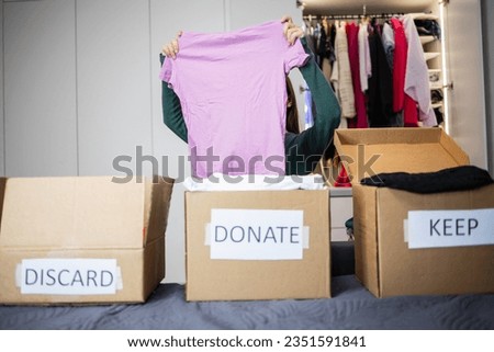 Woman chooses which box to put a pink T-shirt in. Cardboard boxes with labels discard ,keep, donate. Sorting and organizing the storage of clothes, minimalism, thinking about the environment Royalty-Free Stock Photo #2351591841