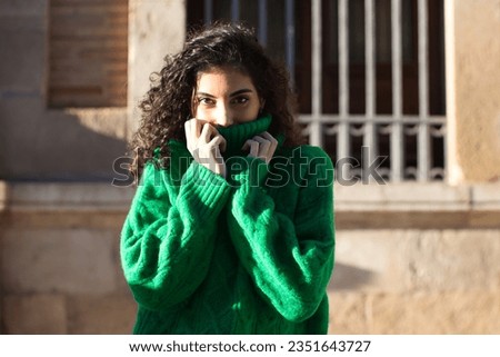 Portrait of beautiful young brunette woman with curly hair and green woollen coat covering her face with the collar of the coat for the cold. The woman is happy and looks at the camera for the photo.