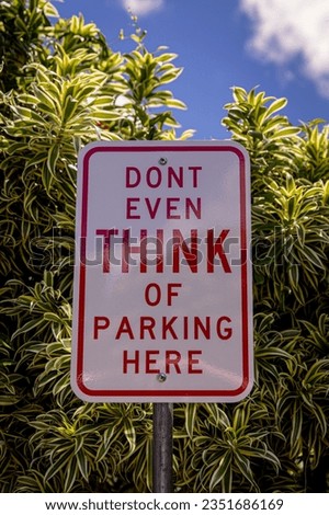 A funny Don't Even Think Of Parking Here Parking Sign located at a parking lot, green bush in the background on a sunny day