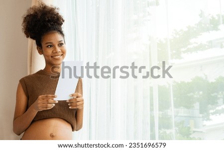 Happy portrait african american pregnant mother standing by home window looking at upcoming baby's ultrasound with fanciful anticipation and delighted to see healthy fetal evolution.