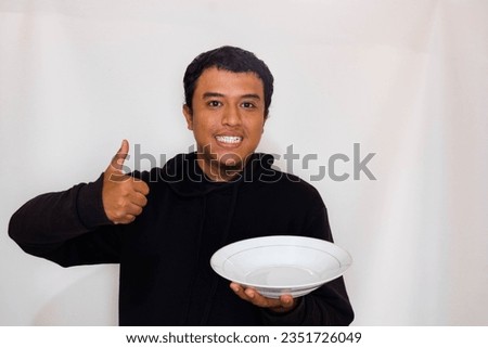 Adult Asian man smiling happy and give thumb up wearing black sweater while showing empty dinner plate.