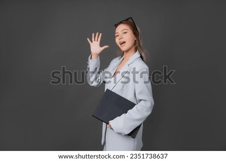 Asian long-haired female executive or company employee Wearing a blue suit and fashion glasses, holding a laptop, raising his hand to form the number 5, taking pictures in a gray background studio.