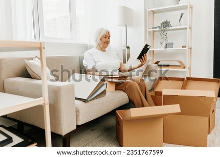 elderly woman sits on a sofa at home with boxes. collecting things with memories albums with photos and photo frames moving to a new place cleaning things and a happy smile.