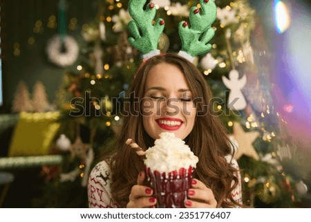 Christmas time. smiling elegant woman with festive hot chocolate cocktail and funny reindeer antlers headpiece hoop in traditional Christmas sweater near Christmas tree at modern home. Royalty-Free Stock Photo #2351776407