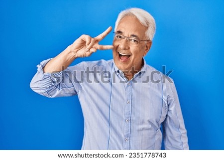 Hispanic senior man wearing glasses doing peace symbol with fingers over face, smiling cheerful showing victory 
