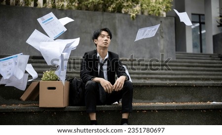 A depressed and sad young Asian businessman is throwing his business paperwork while sitting on the stairs in front of the corporate office with a box of his personal stuff after losing his job. Royalty-Free Stock Photo #2351780697
