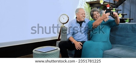 Indian happy old senior couple sitting on comfy sofa taking funny faces selfie using mobile phone at indoor home. Smiling aged husband wife looking at camera posing for photo vlogger recording video