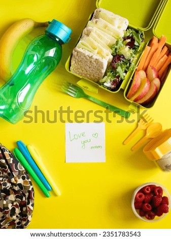 School lunch box with sandwiches, carrot sticks, apple, banana, lettuce, hummus and raspberries. Healthy school lunch concept. Yellow background and note with text - love you mom. Top view 
