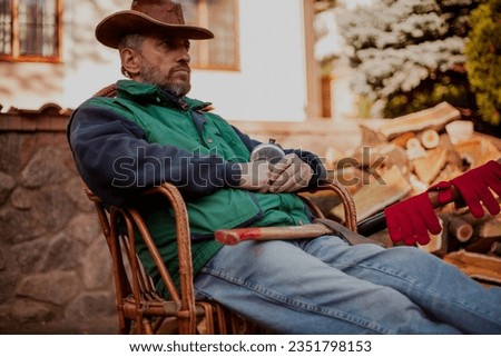 man in a hat is resting in a wicker chair in the yard in front of a pile of chopped wood. He holds a round metal flask in his hands. There is an ax on my feet

