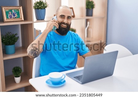 Young bald man using laptop talking on smartphone at home