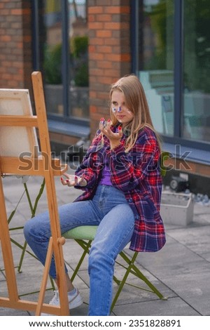 Focused girl artist with paint on her face painting a picture standing on an easel with paintbrush