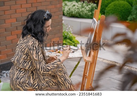 young pretty girl artist sits on a chair in the street and with a brush paints a picture on canvas standing on an easel
