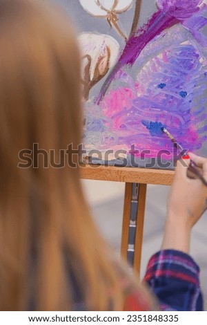 A young girl artist paints picture with a brush on a canvas that stands on an easel view from behind the back
