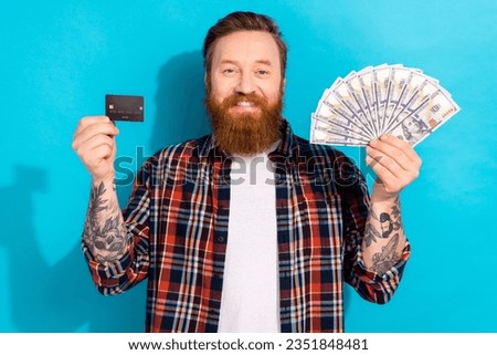 Portrait of cheerful nice man with tattoo wear plaid shirt hold credit card dollars receive payment isolated on blue color background