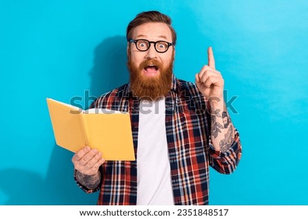 Portrait of modern young teacher with tattoo wear plaid shirt holding book raising finger up have idea isolated on blue color background