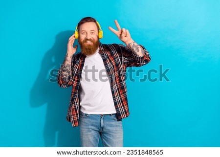 Photo of optimistic man with stylish beard dressed checkered shirt hand touch headphones show v-sign isolated on blue color background